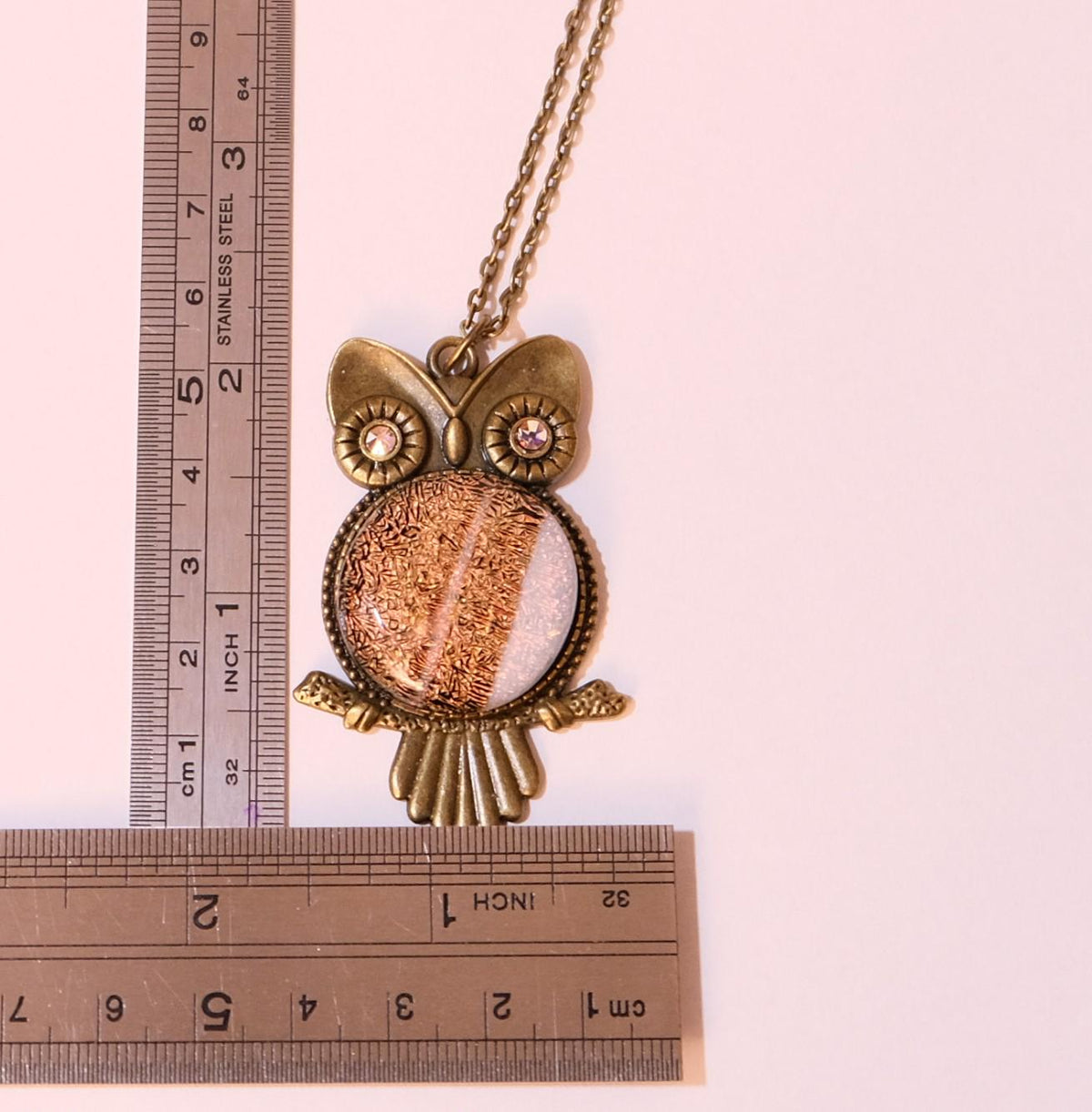 An owl pendant necklace with a bronze-finished metal frame and chain, featuring a textured metallic center and small rhinestone eyes, shown next to metal rulers for scale on a pale pink background.
