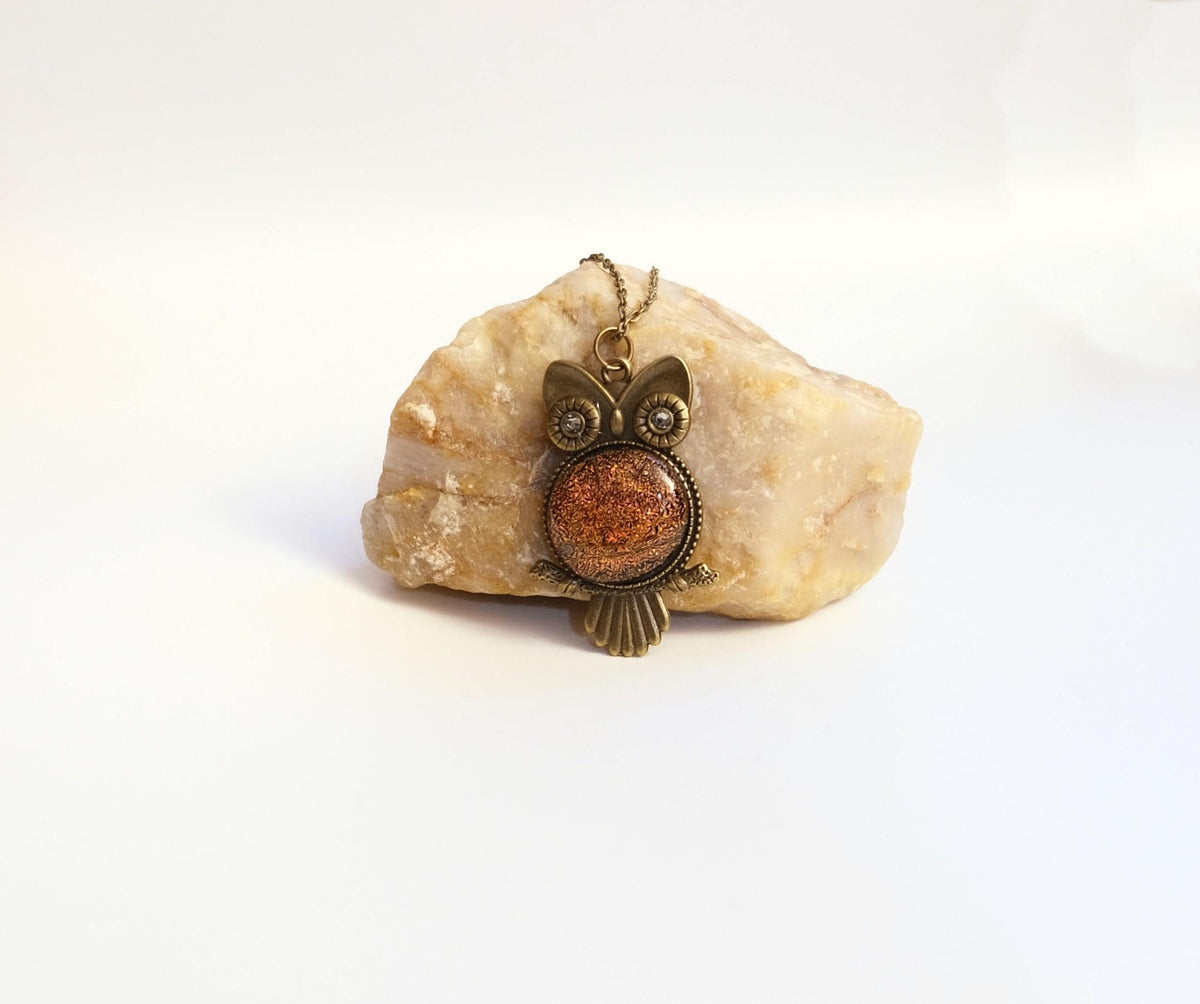 An owl-shaped pendant necklace with a textured copper-orange center and antique bronze metal details, displayed on a light beige raw stone against a white background.