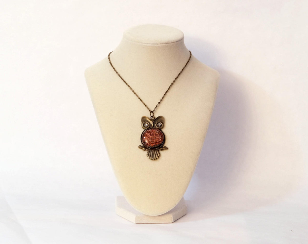 An owl-shaped pendant necklace with a textured copper-orange center and antique bronze metal details, displayed on a light beige raw stone against a white background.