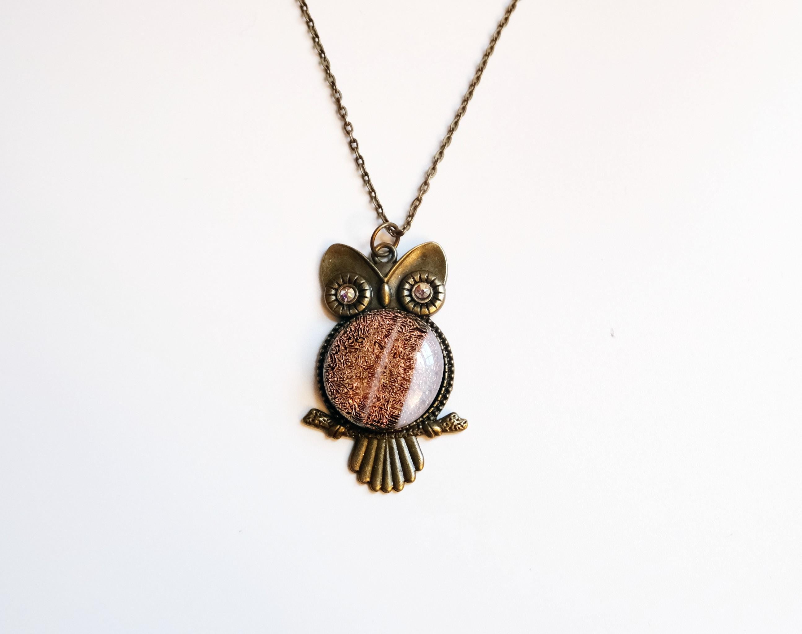 An owl pendant necklace with an antique bronze metal finish and a central oval cabochon showing a pinkish-brown glittery pattern, displayed on a clean white background.