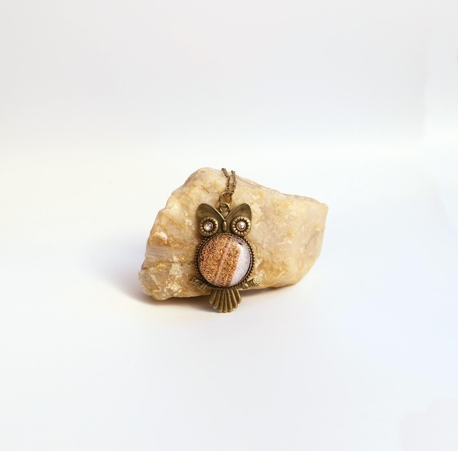 An owl-shaped pendant with a textured brown and white face, set in an antique bronze-tone metal, displayed on a small beige stone against a white background.