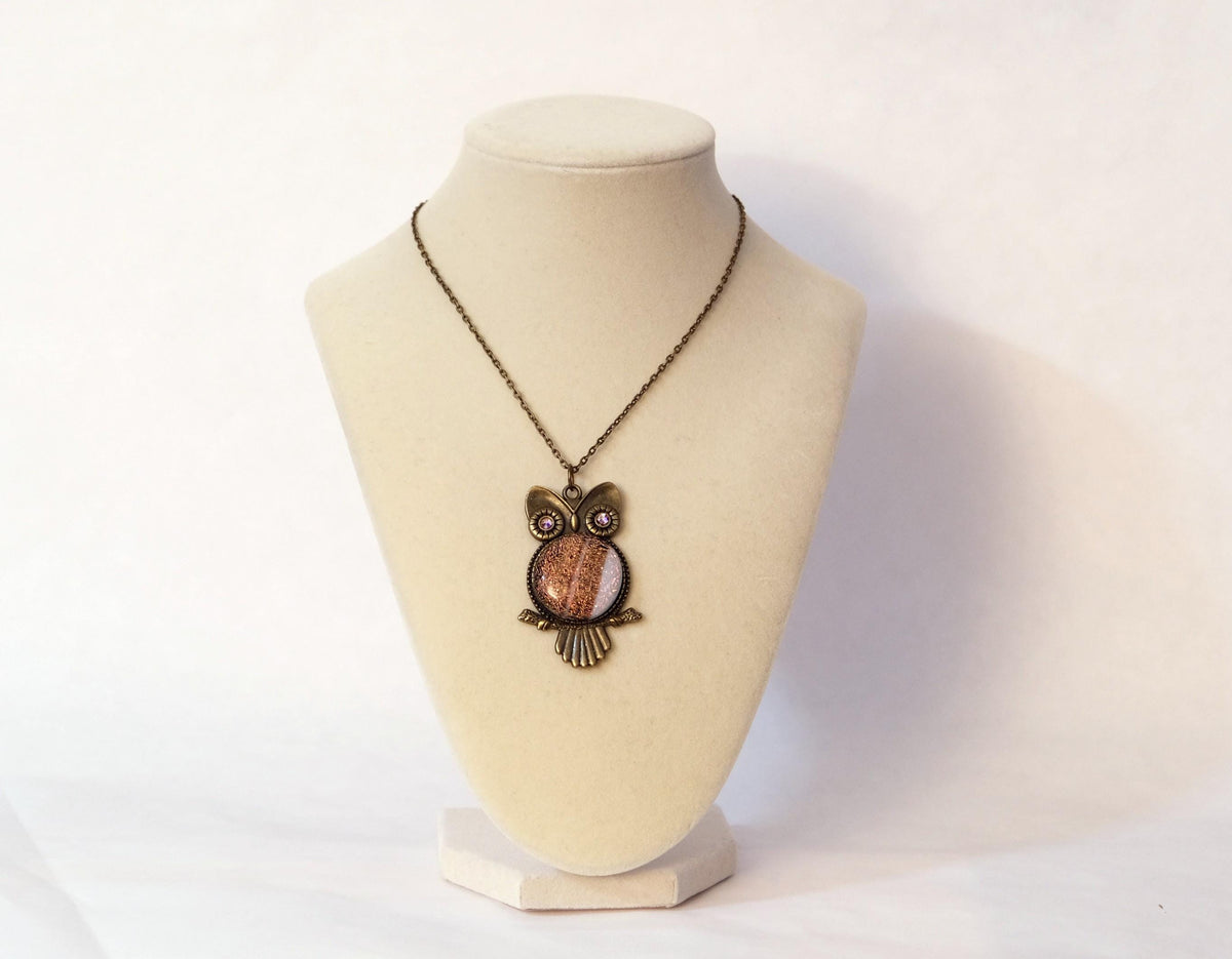 An owl pendant necklace with a copper and dark brown metal finish, displayed on a cream-colored jewelry bust against a white background.