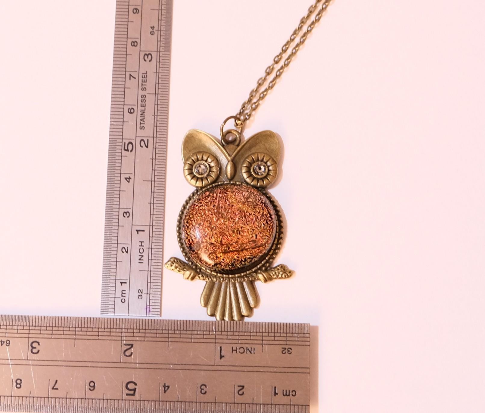 An owl-shaped pendant necklace with a textured copper-orange center and antique bronze metal details, displayed on a light beige raw stone against a white background.