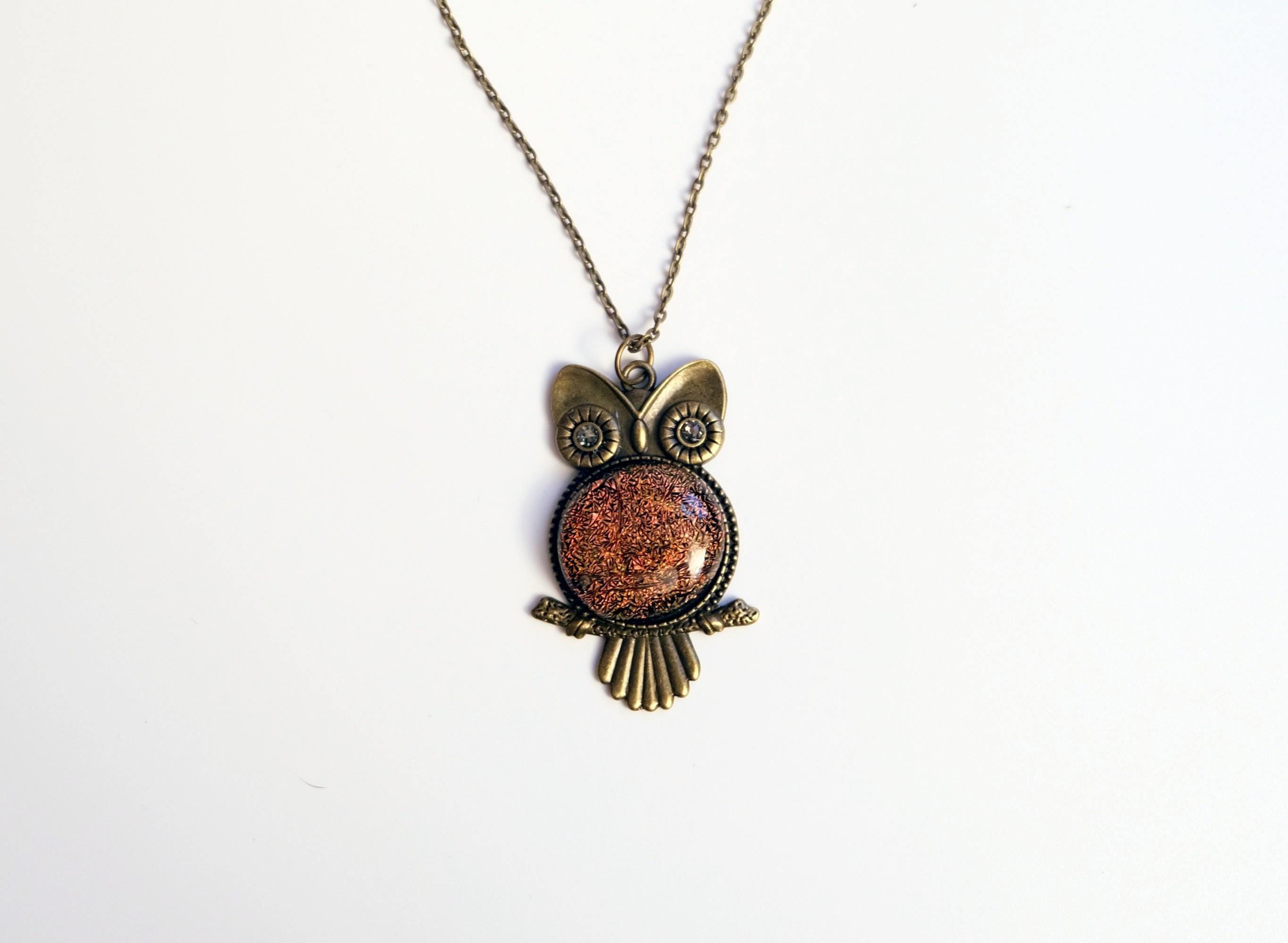 An owl-shaped pendant necklace with a textured copper-orange center and antique bronze metal details, displayed on a light beige raw stone against a white background.