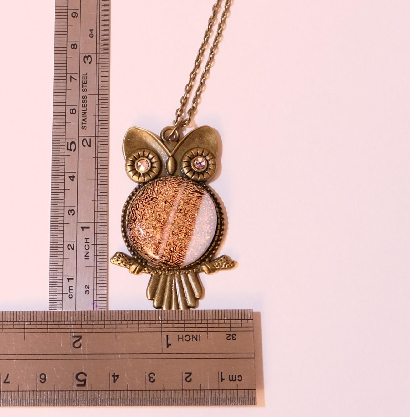 An owl pendant necklace with a bronze-finished metal frame and chain, featuring a textured metallic center and small rhinestone eyes, shown next to metal rulers for scale on a pale pink background.
