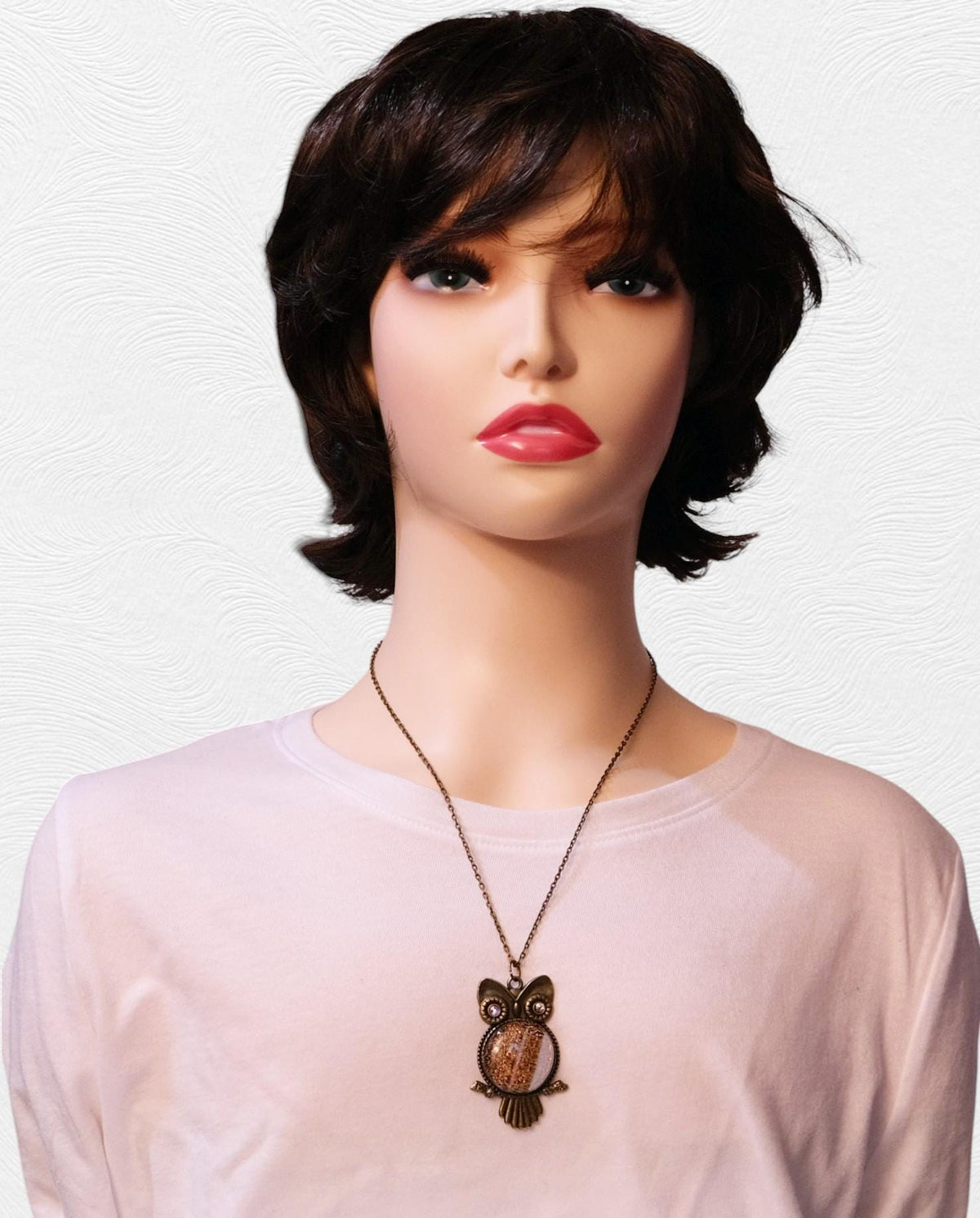 A mannequin wearing a short dark brown wig with bangs, a pale pink long-sleeve top, and an owl pendant necklace against a light textured background.
